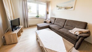 Ein Wohnzimmer mit braunem Sofa, einem weißen Tisch mit Spitzenleinentuch, einem Holz-TV-Schrank mit Flachbildfernseher und einem Fenster mit Vorhängen.