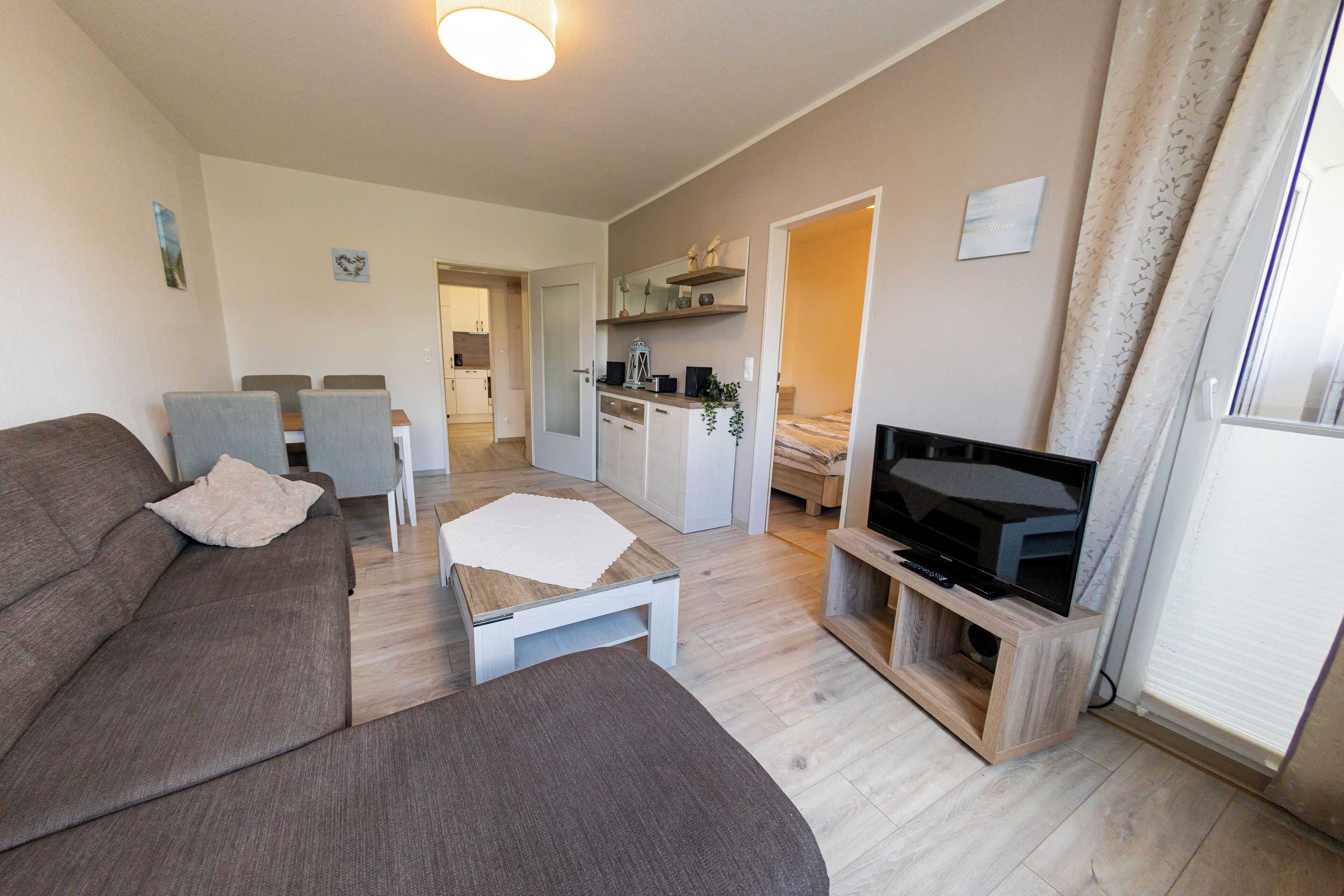 A modern living room with a sofa, a coffee table, and a TV on a wooden stand. There is a dining area with chairs and a table, and a kitchen in the background.