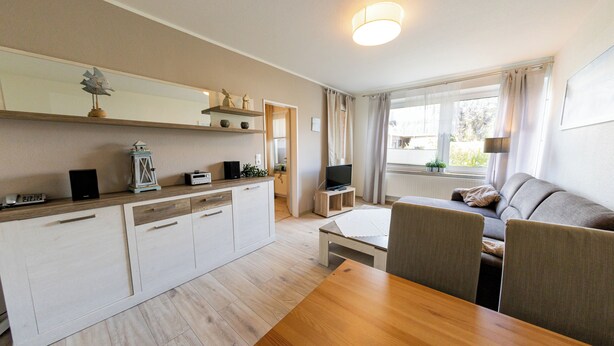 Ein modernes Wohnzimmer mit Esstisch aus Holz, grauem Sofa und Küchenbereich mit weißen Schränken und einem integrierten Fernseher.