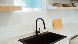 A kitchen with a black sink, white marble countertop, wooden shelves, and a window with a view of trees. - Adorable Studio - Private Patio - Dream Kitchen (Waco)