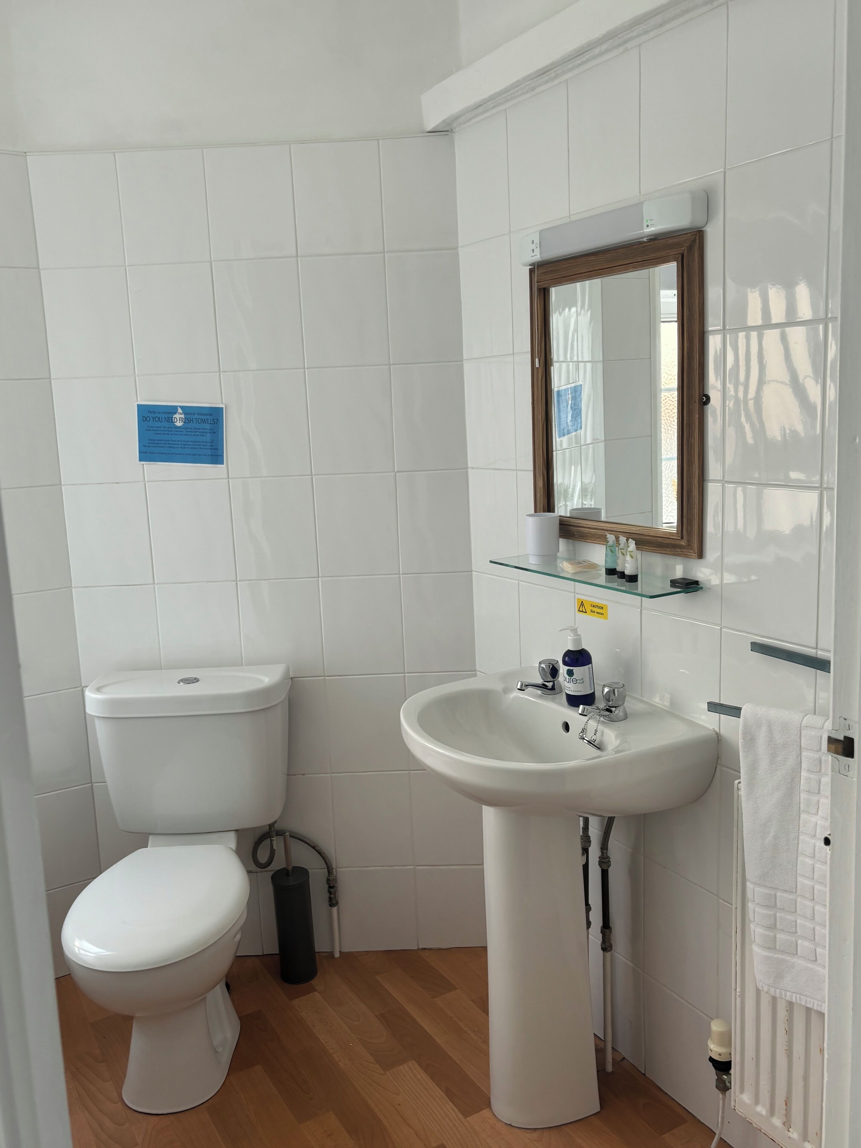A small, clean bathroom with white tiles, a toilet, a sink with a mirror above it, and a glass shelf.