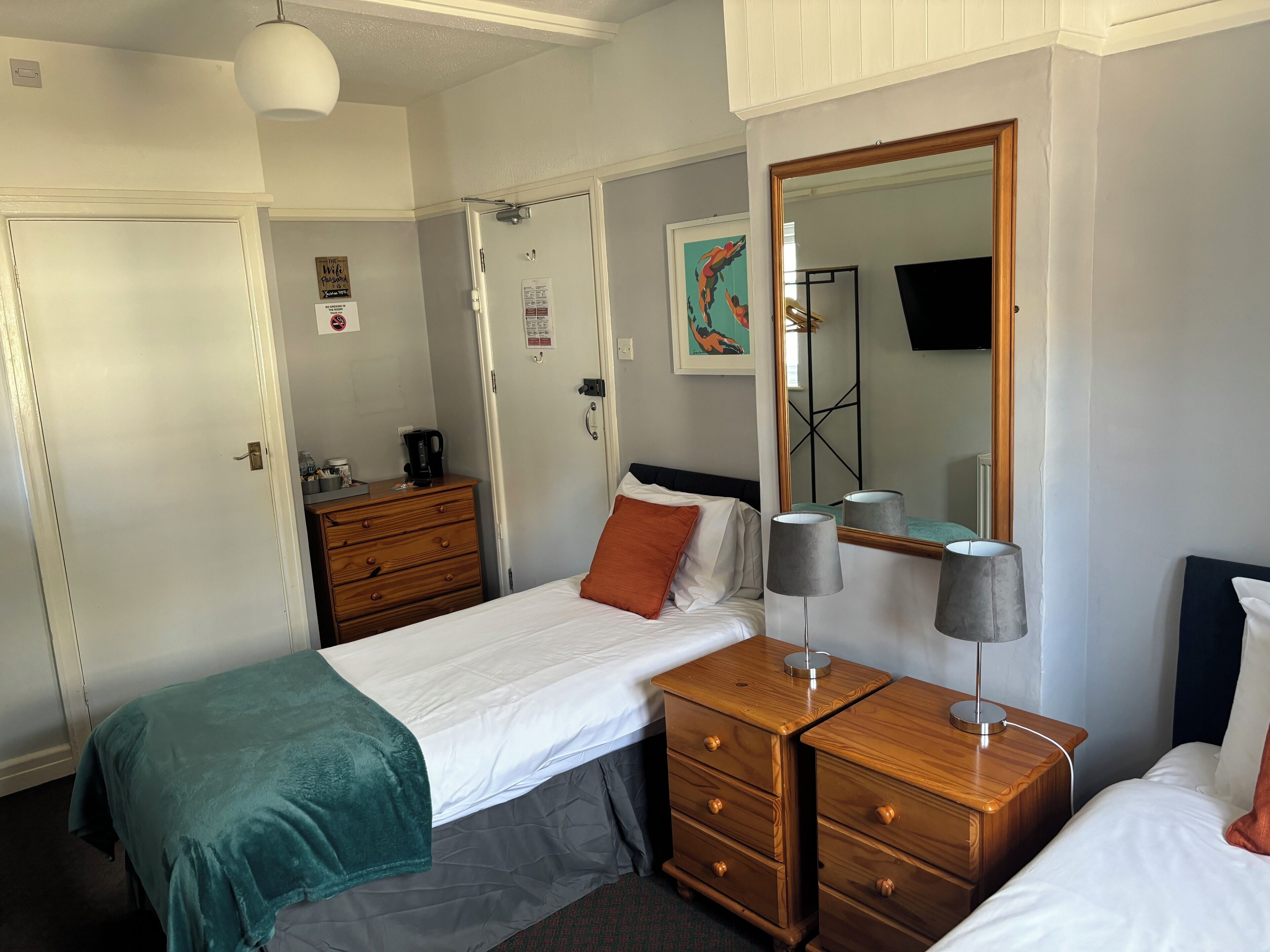 A hotel room with a bed, a wooden dresser, a desk with a lamp, a mirror, and a television.