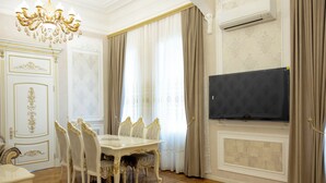 A dining room with a white table and chairs, a chandelier, and a flat-screen TV mounted on the wall.