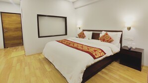 Habitación de hotel con piso de madera, cama con ropa blanca y un plaid rojo estampado, mesita de noche con artículos de tocador y un televisor montado en la pared.