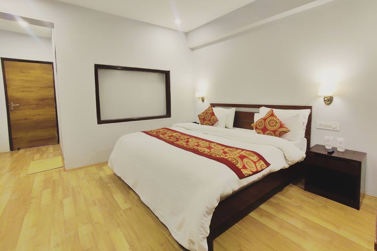 A hotel room with a wooden floor, a bed with white bedding and red patterned throw, a nightstand with toiletries, and a wall-mounted TV.