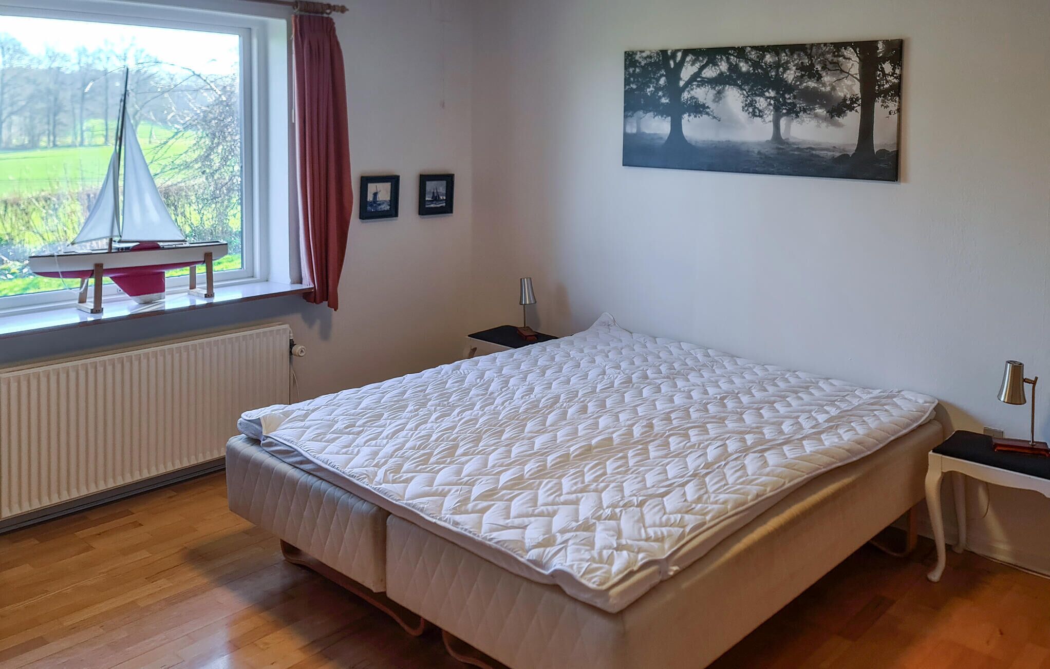Ein Schlafzimmer mit einem großen Bett, einem Fenster mit Blick auf Bäume, einem Heizkörper und einem kleinen Tisch mit einer Lampe.