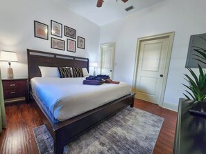A neatly arranged bedroom with a large bed, bedside tables, a nightstand, a door, and a wall with framed artwork.