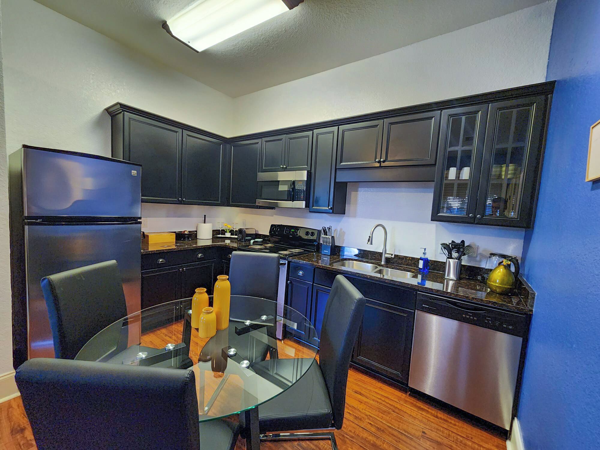 A modern kitchen with dark cabinetry, stainless steel appliances, and a glass dining table.