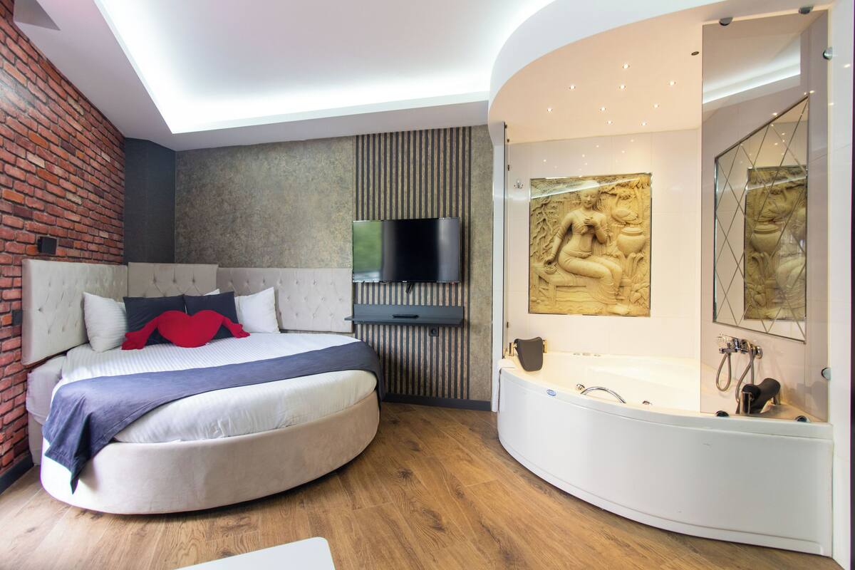 A modern bedroom with a curved bed, a built-in TV, a bathtub, and a stone wall.
