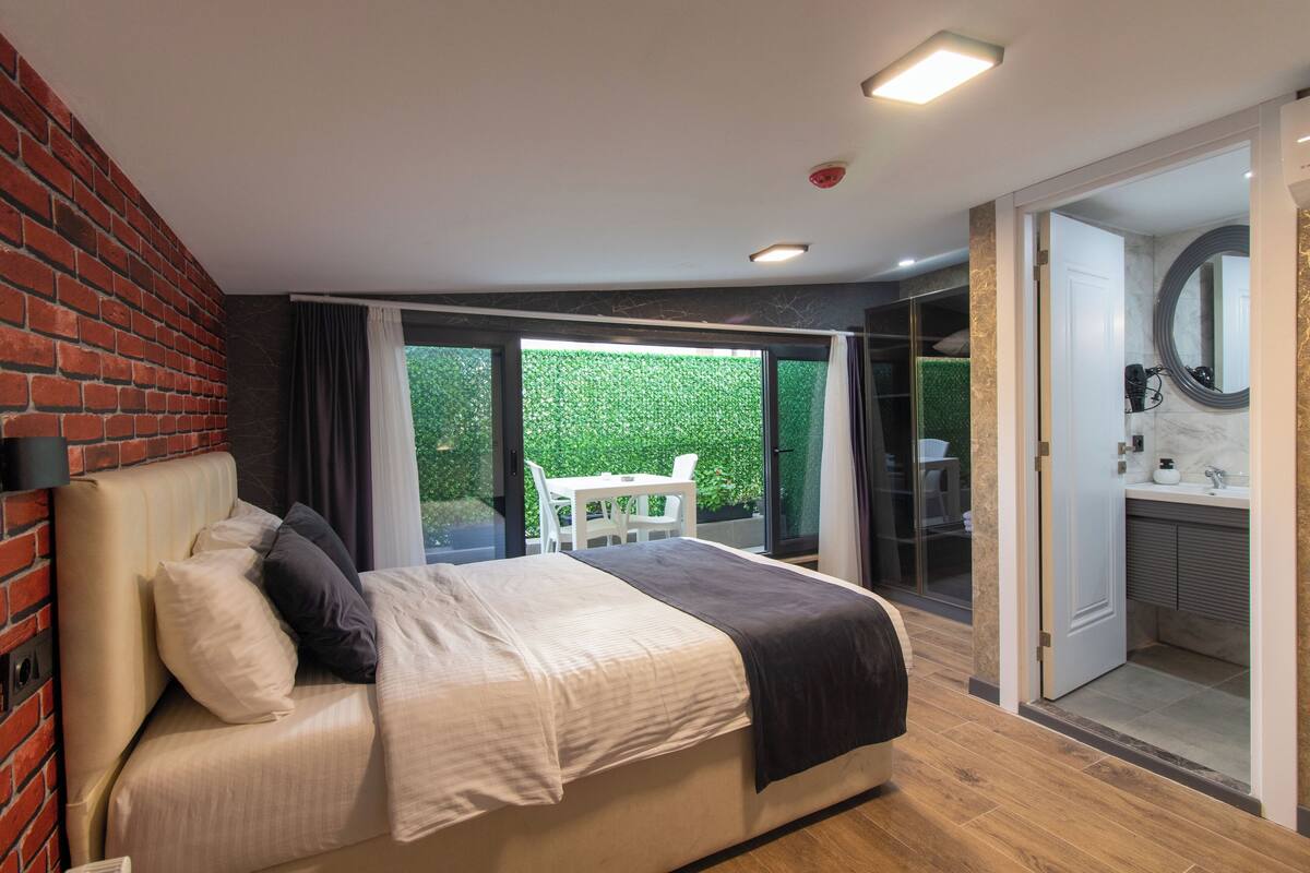 A bedroom with a bed, a white chair, and a view of a green garden through a sliding glass door.