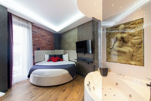 A modern bedroom with a curved bed, a large window with curtains, a flat-screen TV, a wall-mounted TV, a bathtub, and a decorative wall piece.