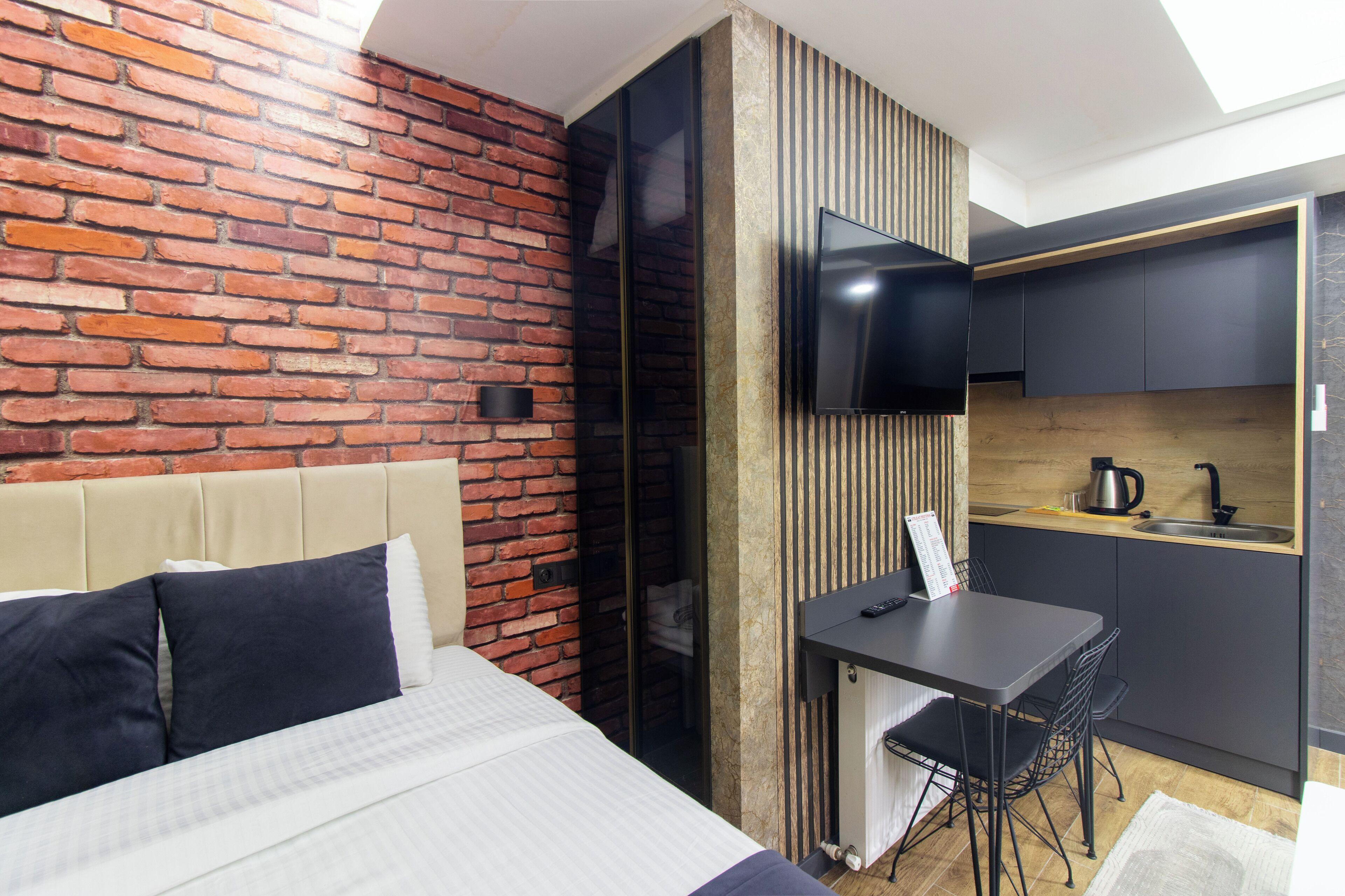 A hotel room with a brick wall, a bed with white and dark blue bedding, a small desk with a chair, a television, and a kitchenette with a sink and a kettle.