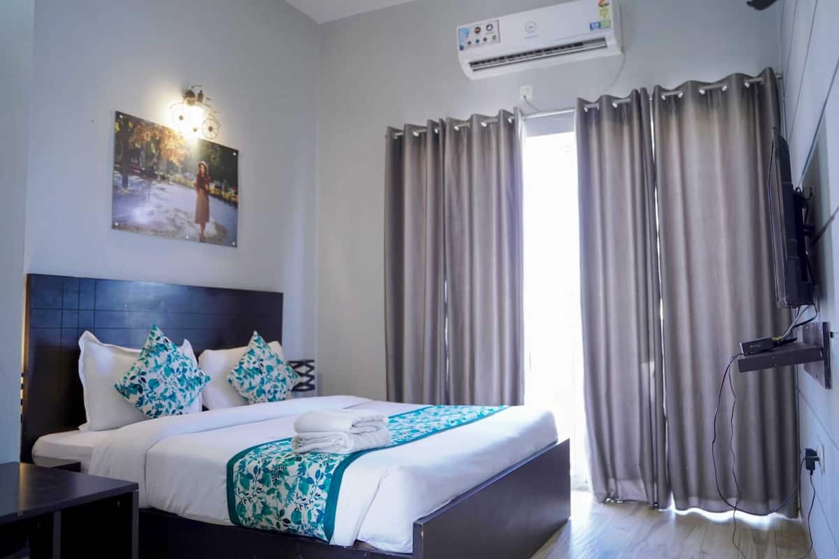 A hotel room with a bed, a wall-mounted air conditioner, a painting, and a television.