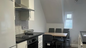 Een kleine keuken met een kookplaat, oven en witte kastjes. Er staat een eettafel met stoelen voor een raam.