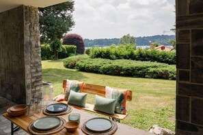Patio con tavolo in legno apparecchiato per la cena, una panca con cuscini e vista su un giardino curato e uno specchio d'acqua.