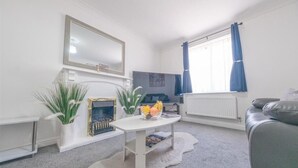 Un salon moderne avec un canapé blanc, une table basse sur laquelle sont disposés des fruits, un téléviseur et un grand miroir.