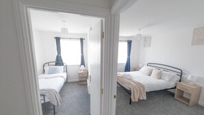 Une chambre avec deux lits, une table de chevet, une commode, une fenêtre avec des rideaux et une porte donnant sur une autre pièce.