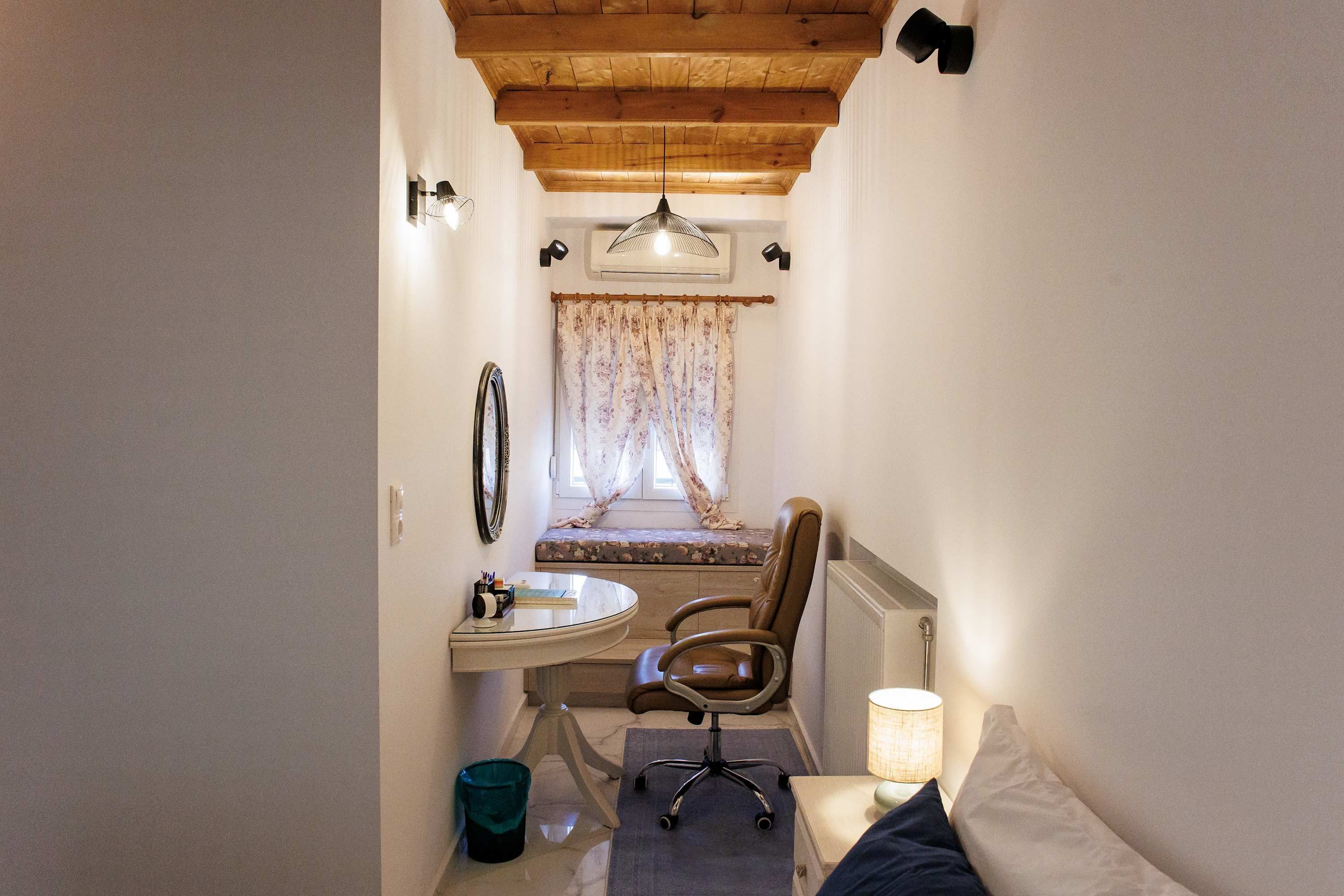 A narrow hallway with a wooden ceiling, a bench with a floral curtain, a desk with a sink, a chair, and a bedside lamp.
