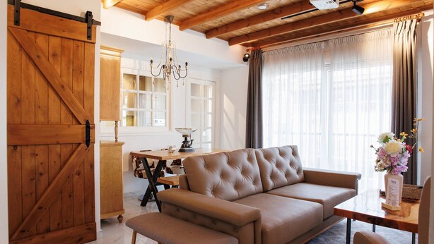 Una sala de estar acogedora con sofá, banca, techo de madera, candelabro y ventana con cortinas.