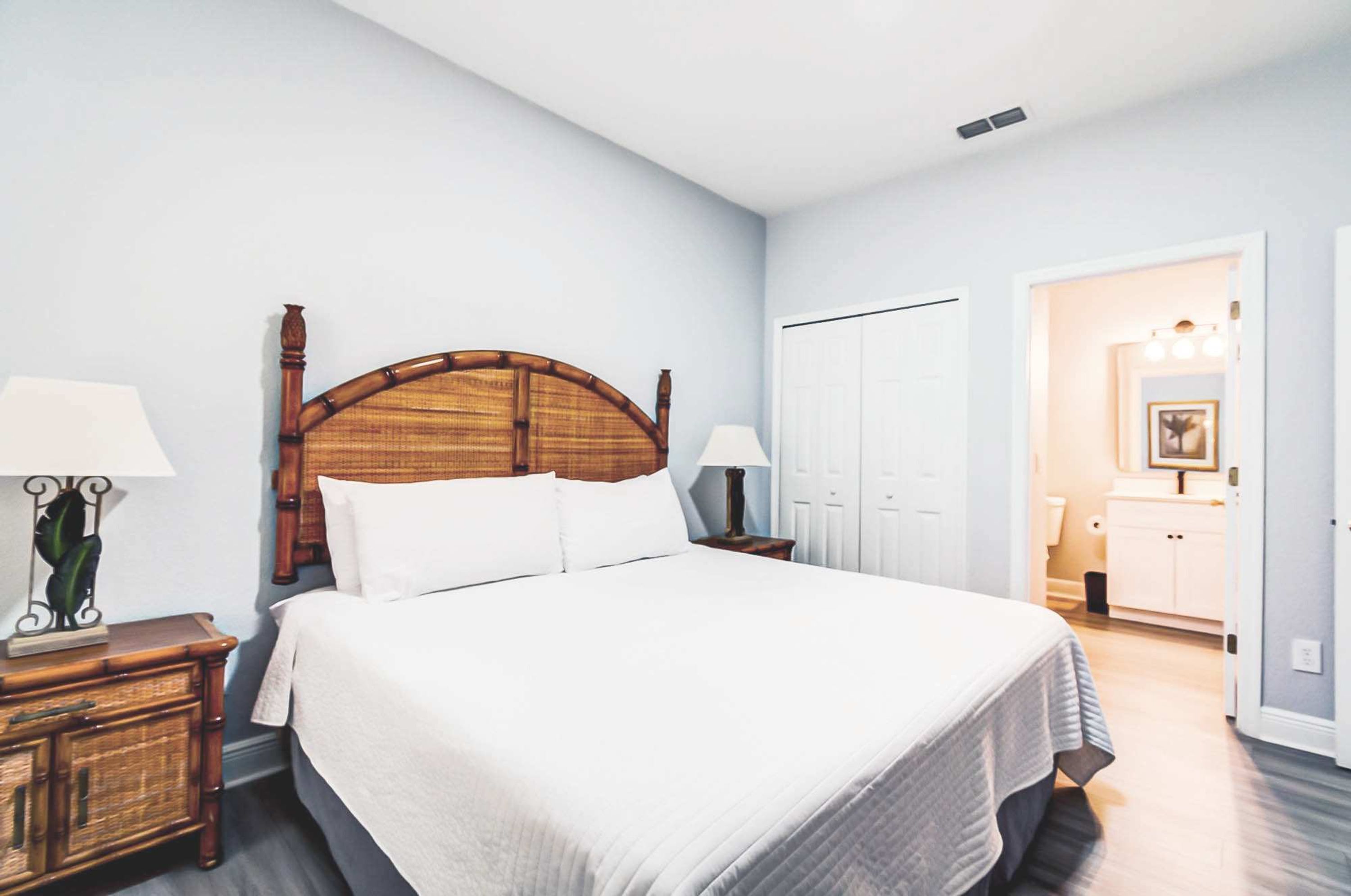 Quarto com cama grande, cabeceira de madeira, duas mesas de cabeceira com luminárias, guarda-roupa e banheiro visível por uma porta aberta.