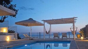 Ein Swimmingpool mit freiem Blick auf das Meer, eine Pergola mit Sonnenschirmen und Liegestühlen.