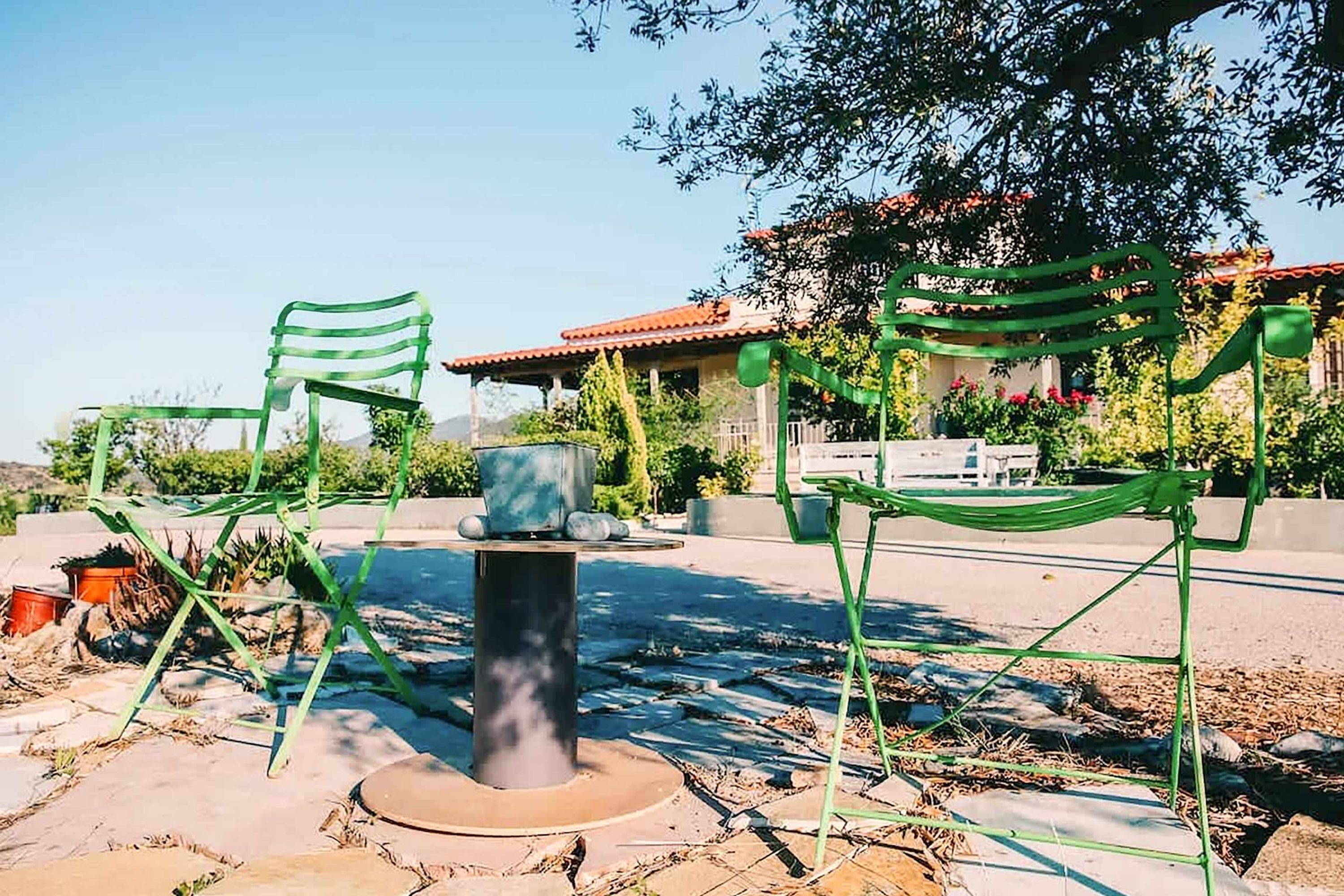 Mobiliario exterior con sillas de metal y una mesa, rodeado de plantas y un edificio con techo de tejas rojas.