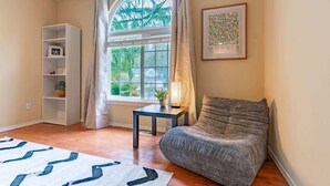 A cozy living room with a large window, a bookshelf, a small table with a lamp, and a comfortable sofa.