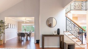 A spacious living area with a dining table, chairs, a console table with a mirror, and a staircase with a wrought iron railing.