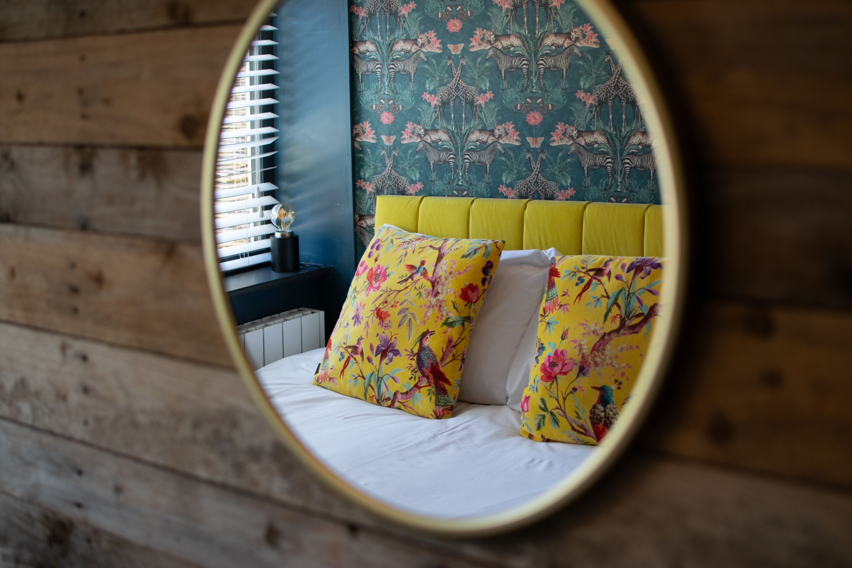 Una cama con almohadones florales, cabecera amarilla y papel tapiz con motivos naturales.