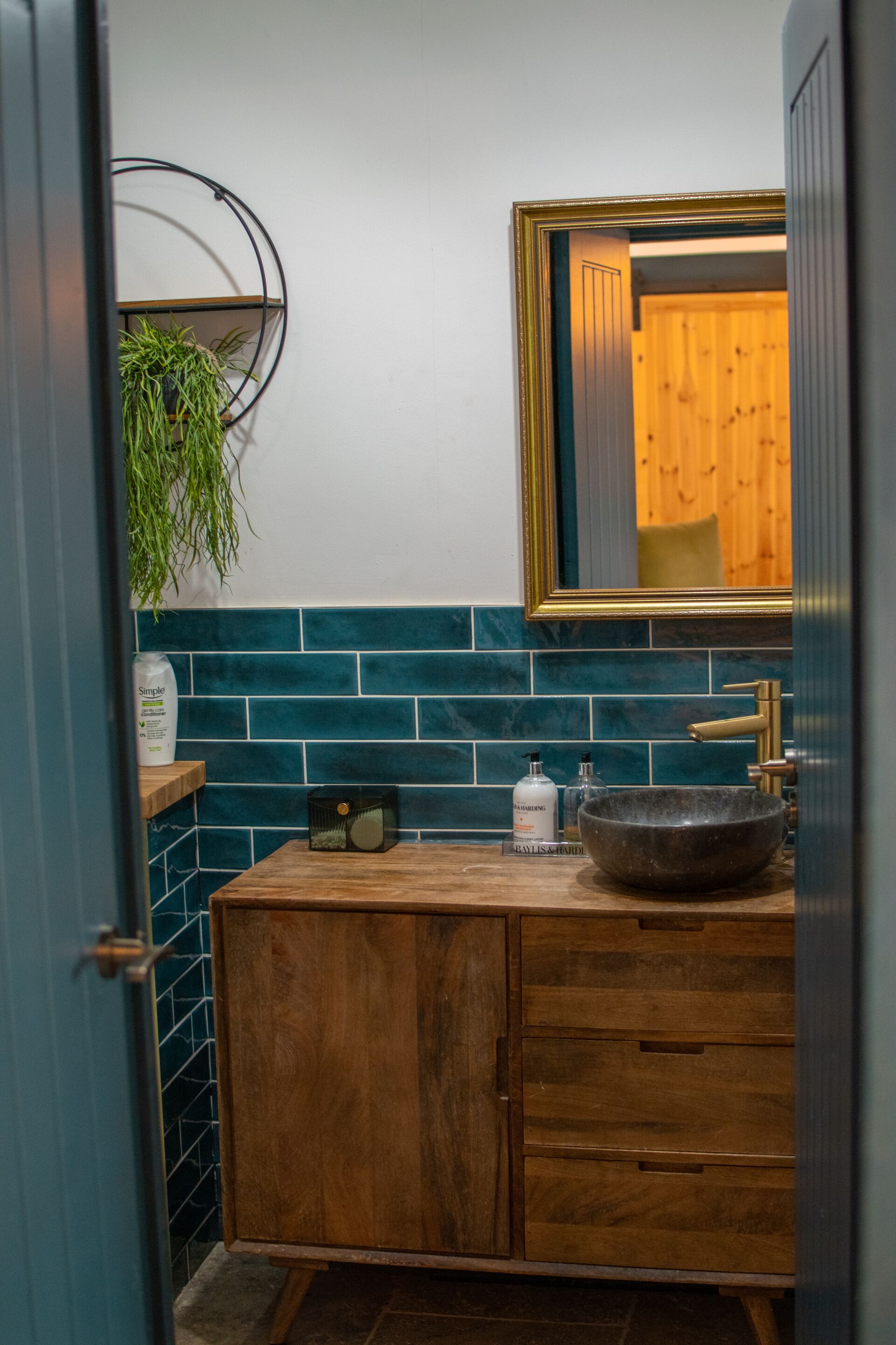 A bathroom with a wooden vanity, a large bowl sink, a mirror with a gold frame, and a green tiled backsplash.