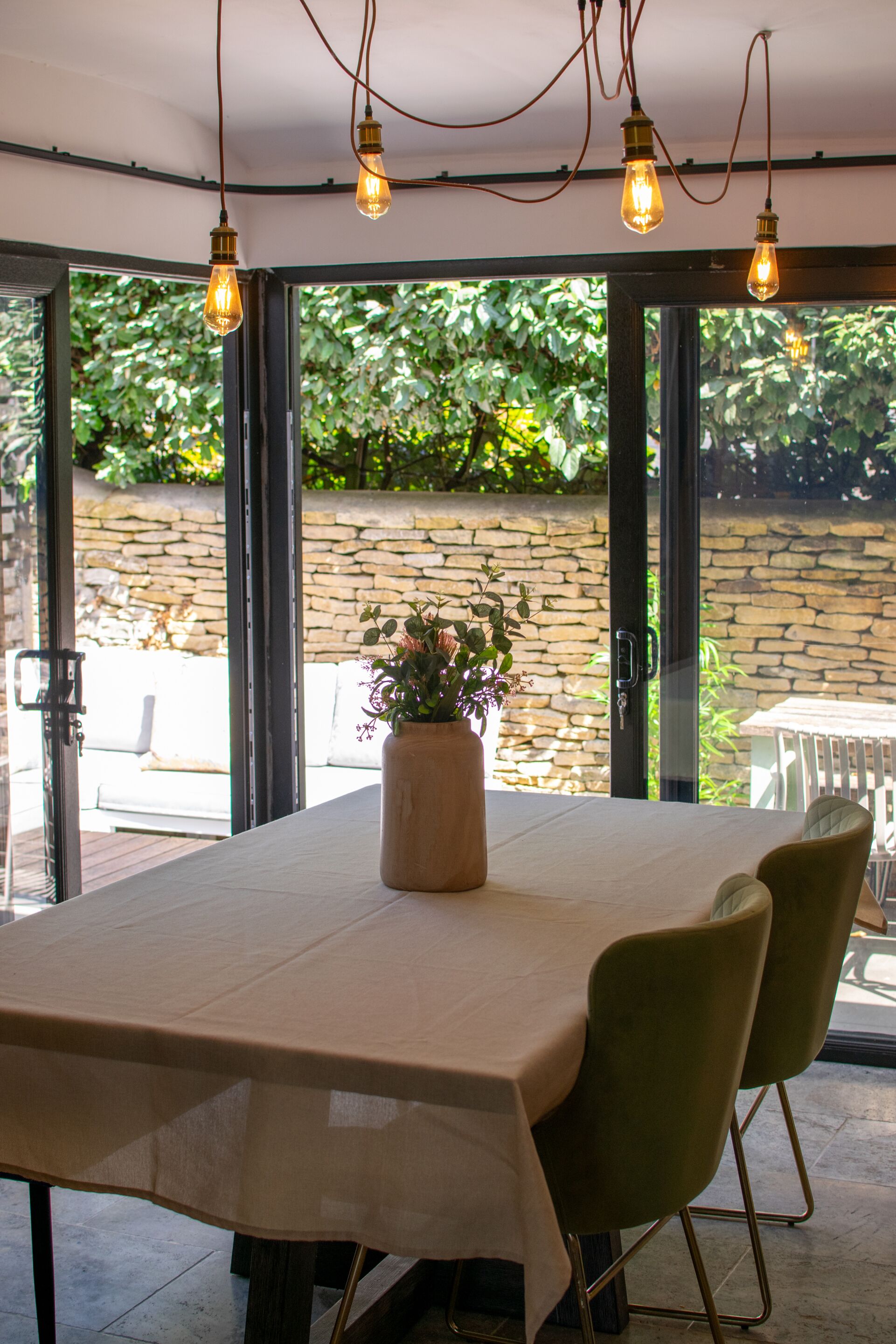 A dining area with a table, chairs, a vase with flowers, and a view of greenery through glass doors.
