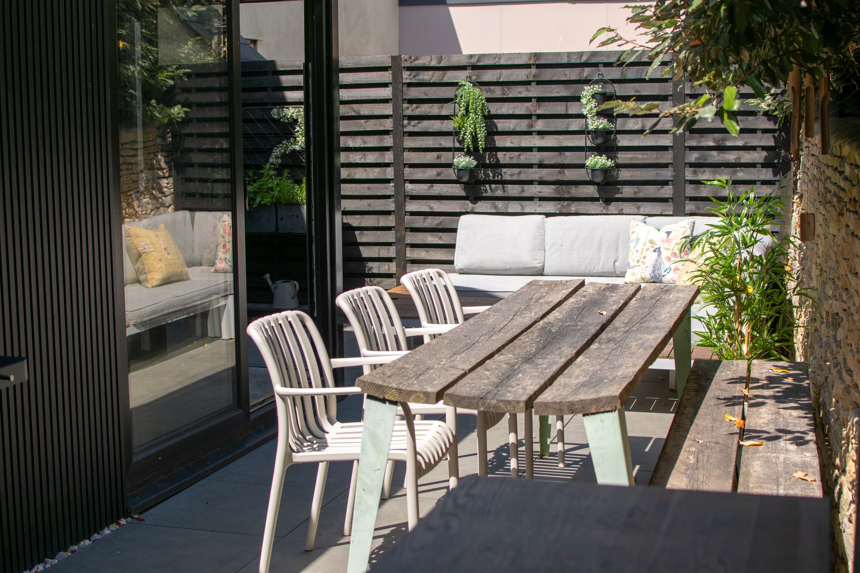 A patio with a wooden table and white chairs, a grey sofa, and a wooden fence with hanging plants.