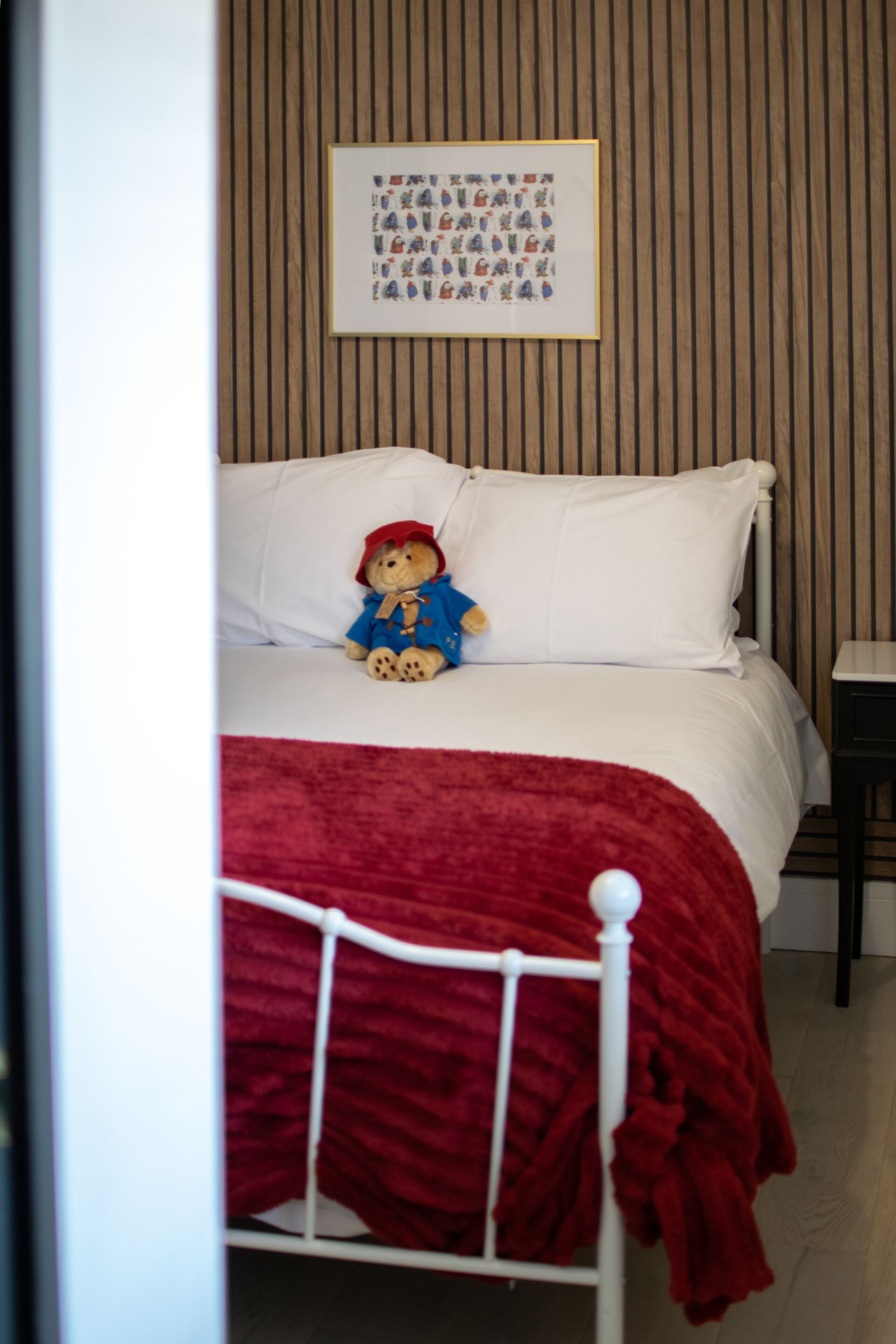 A bed with a teddy bear, a framed picture on the wall, and a red blanket on the floor.