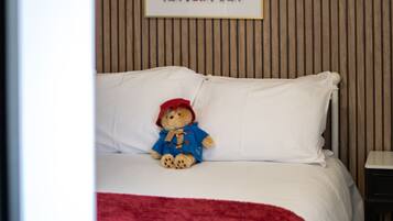 A bed with a teddy bear, a framed picture on the wall, and a red blanket on the floor.