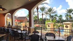 An outdoor dining area with tables and chairs, surrounded by palm trees and a pool.