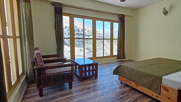 A room with a bed, a wooden chair, a small table, and a large window offering a view of buildings and mountains. - The old monk (Spiti)