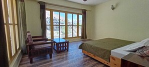 A room with a bed, a wooden chair, a small table, and a large window offering a view of buildings and mountains. - The old monk (Spiti)