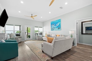 A spacious living room with a grey sofa, teal armchair, and a large painting of dolphins. - Bayfront home with excellent view of the D.I. Bridge (Dauphin Island)