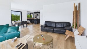A modern living room with a black leather sofa, a teal armchair, and a round coffee table. A dining area with a table and chairs is visible in the background. - Modern house for 8 at 400m from the beach (Plouguerneau)