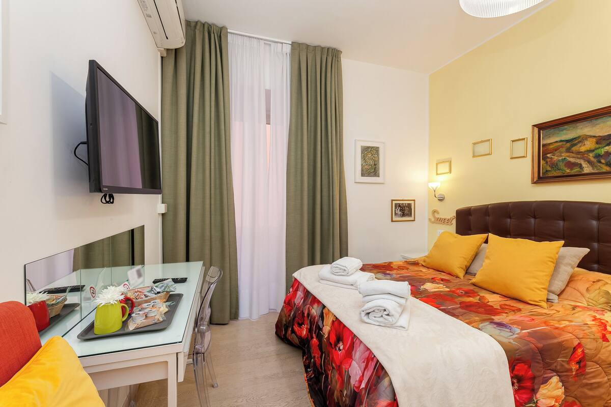 A hotel room with a bed, a TV, a desk with a chair, and a window with curtains.