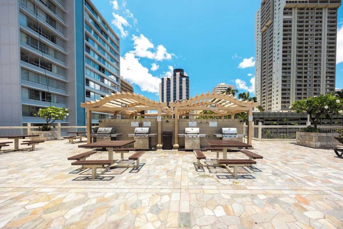 A rooftop patio with barbecue stations, outdoor seating, and a view of surrounding high-rise buildings.