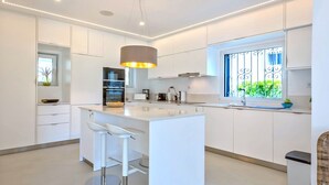 Une cuisine moderne avec des meubles blancs, un îlot central doté de tabourets de bar, un luminaire suspendu et une fenêtre ornée de barreaux décoratifs.