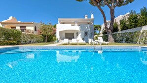 Une piscine avec des chaises longues, un bâtiment blanc doté d’un balcon, et une végétation luxuriante tout autour.