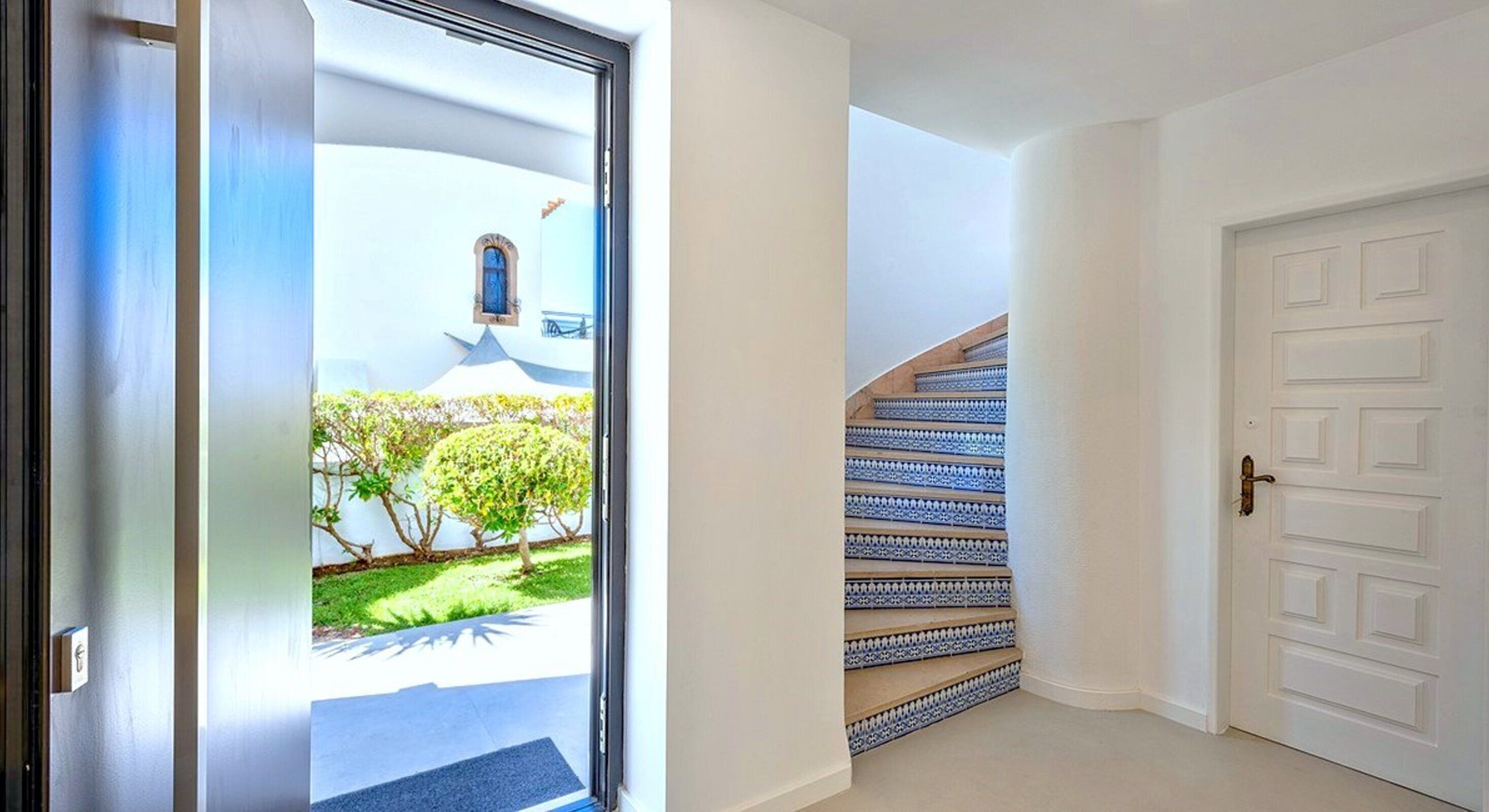 A hallway with a white door, a staircase with blue patterned steps, and a view of a building with a blue facade and greenery outside.