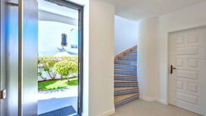Un couloir avec une porte blanche, un escalier dont les marches sont ornées de motifs bleus, et une vue sur un bâtiment à la façade bleue, entouré de verdure.