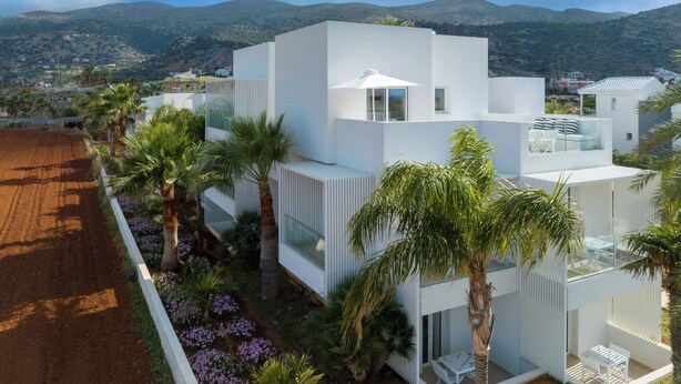 A modern white building with multiple balconies, surrounded by palm trees and purple flowers. - Akrogiali Garden Hotel (Hersonissos)