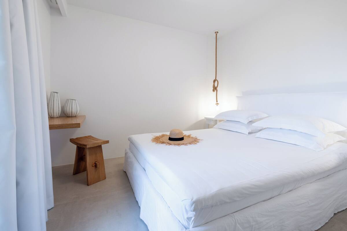 Un dormitorio blanco con una cama, un taburete de madera y dos jarrones en una estantería.