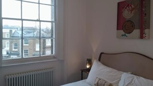 Une chambre dotée d’une grande fenêtre, d’un radiateur, d’un lit avec une literie blanche et d’une œuvre d’art accrochée au mur.