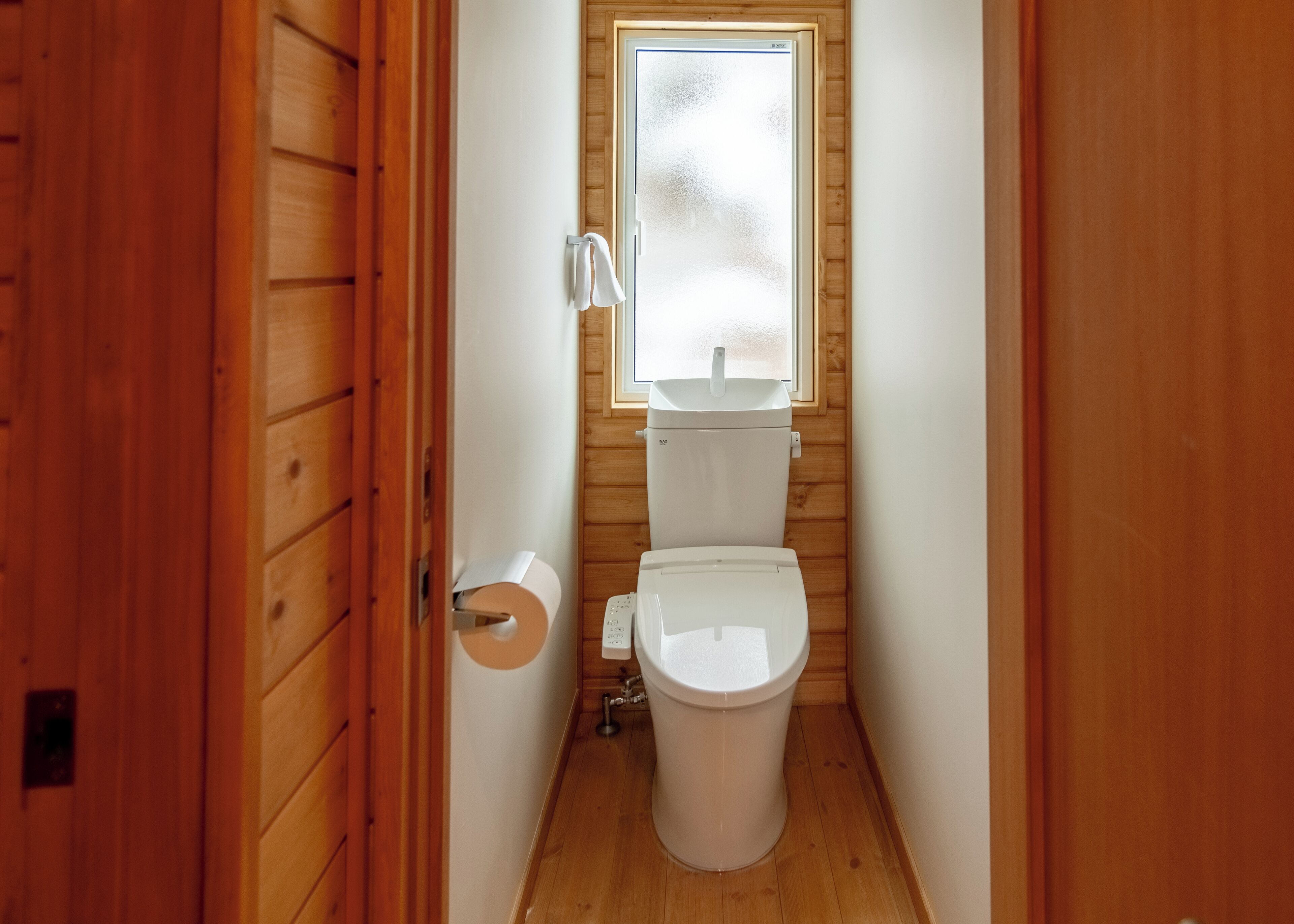 A narrow wooden bathroom with a toilet, a small window, and a towel rack.