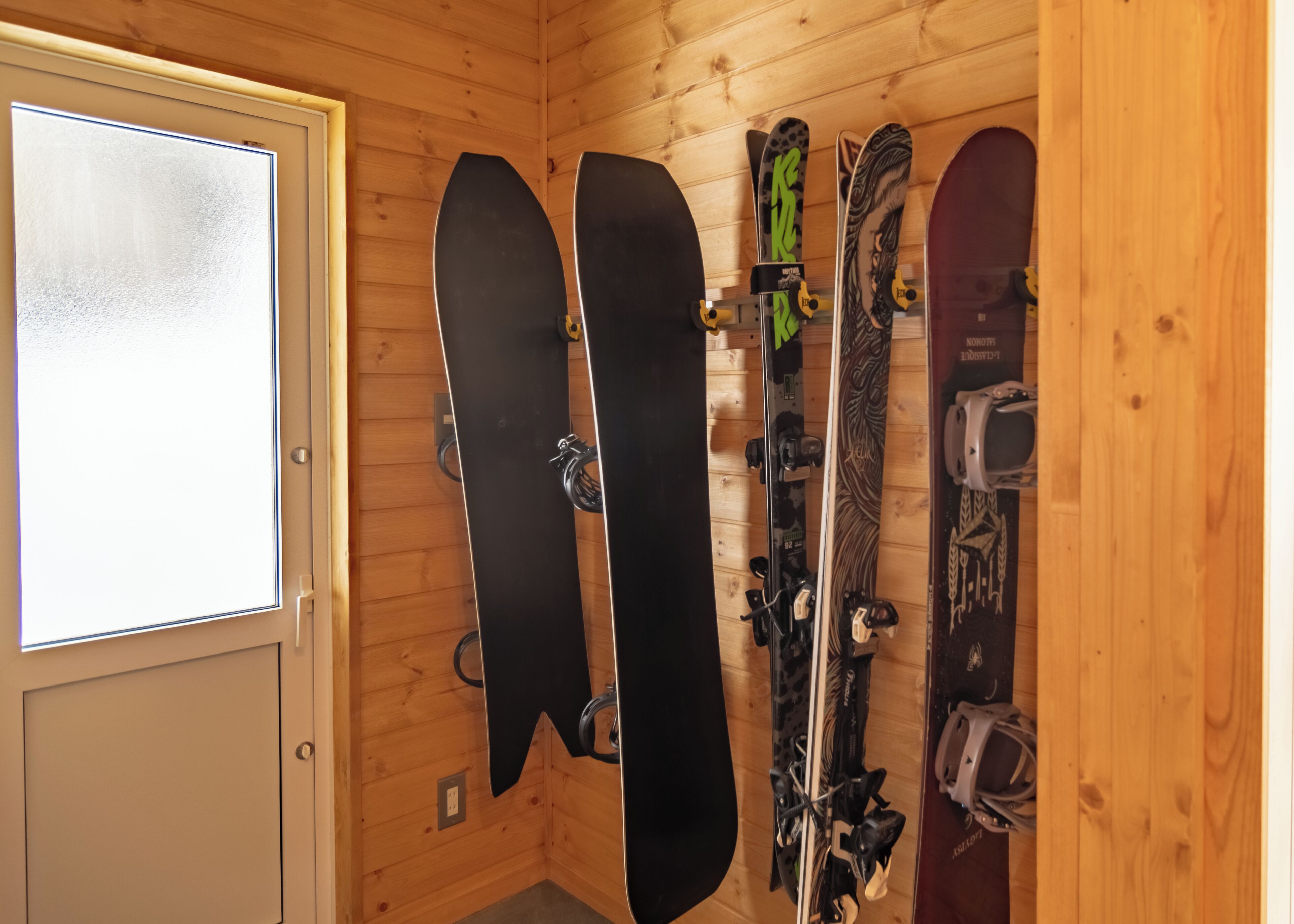 A wooden room with a door, three skis, and a snowboard leaning against the wall.